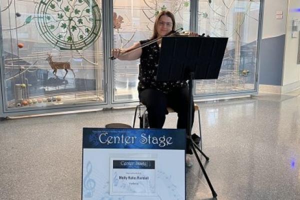 Molly Rahe-Randall is seated in front of a display in the lobby of Penn State Health Milton S. Hershey Medical Center playing a violin. In front of her is a sign that identifies her performance as part of the Center Stage Arts in Health program that provides music in the lobbies of the hospital.